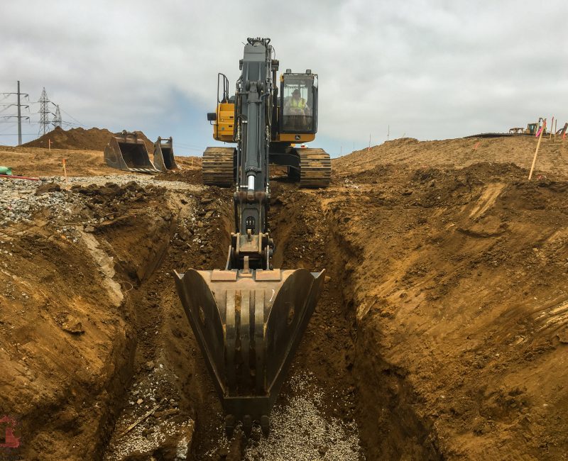 Excavator digs trench at site; operator visible inside cab.