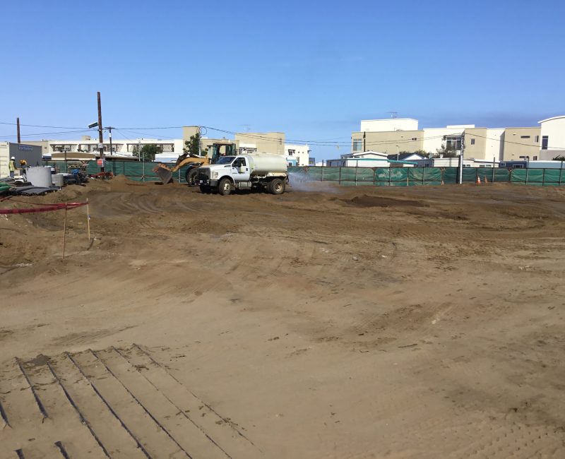 Construction site with water truck, bulldozer, dirt, green fence.