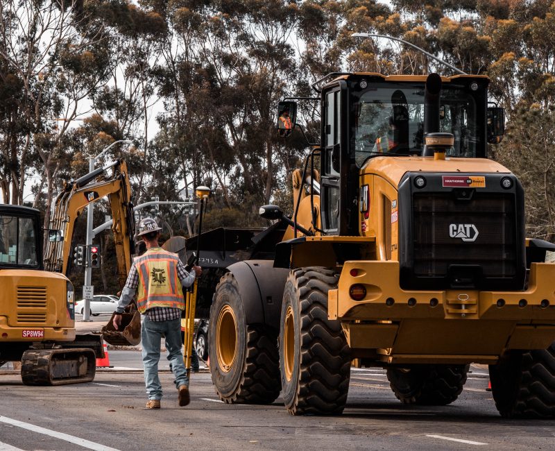 Worker in reflective vest walks beside yellow CAT vehicles on road.
