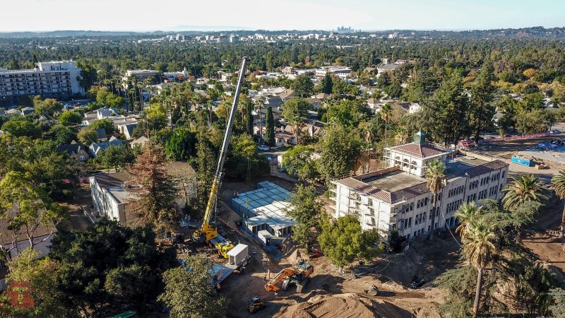 Aerial view: cranes, dirt piles, old buildings, city skyline beyond.