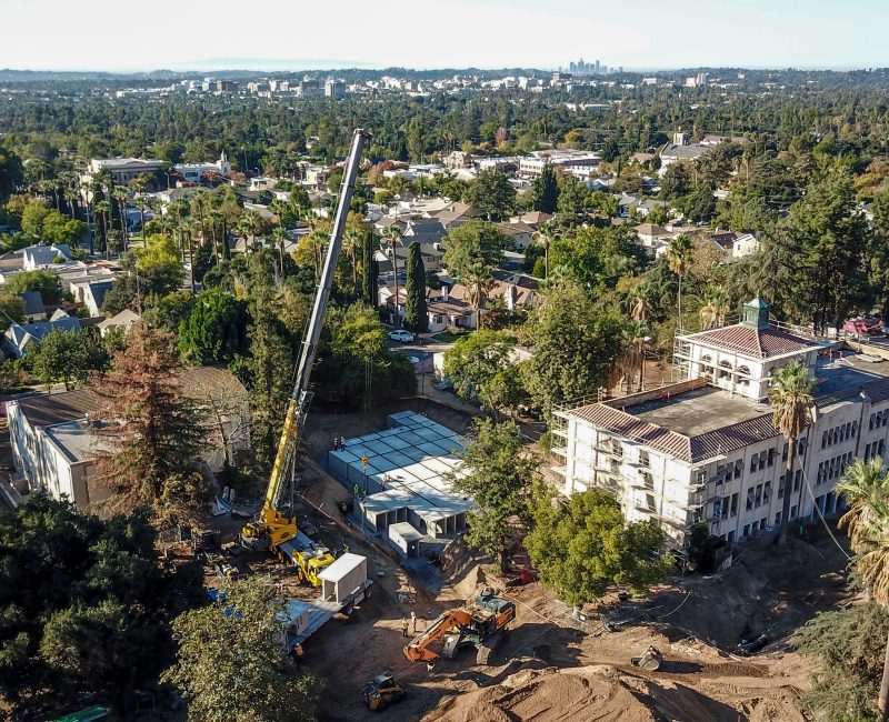Aerial view: cranes, dirt piles, old buildings, city skyline beyond.