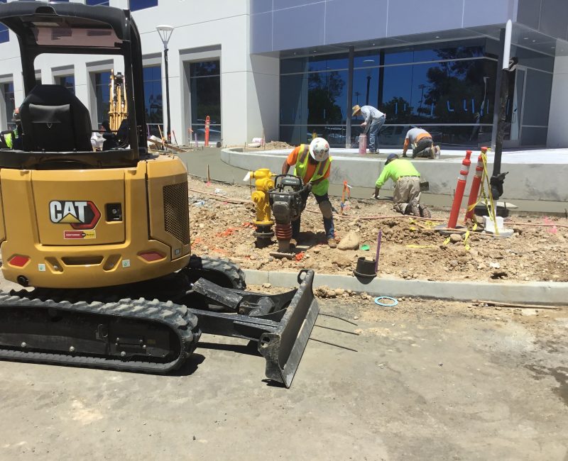 Workers in safety vests by modern building with yellow CAT excavator.