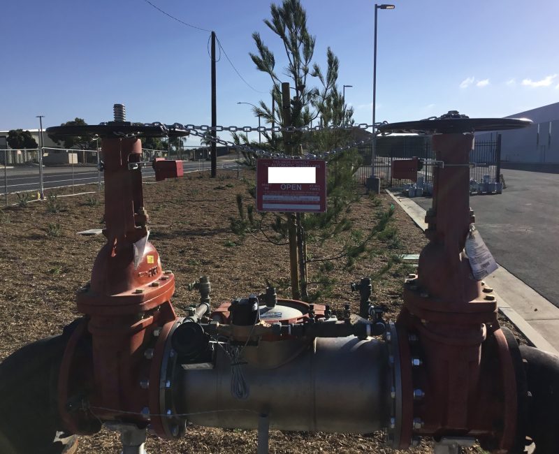 Large red water valve assembly outdoors near tree and sign.