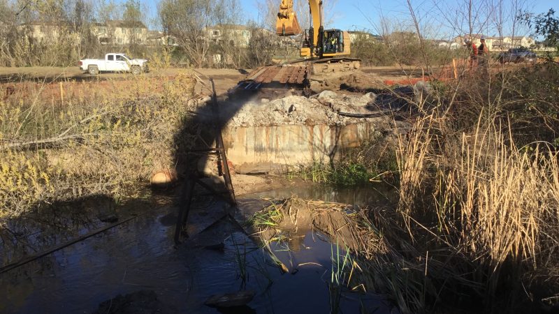 Yellow excavator demolishes a bridge over muddy stream, truck nearby.