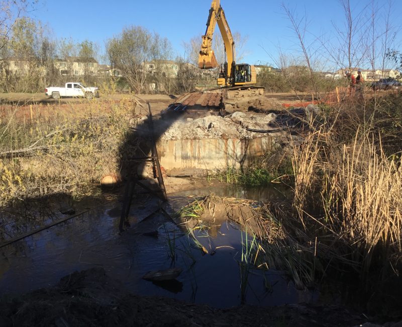 Yellow excavator demolishes a bridge over muddy stream, truck nearby.