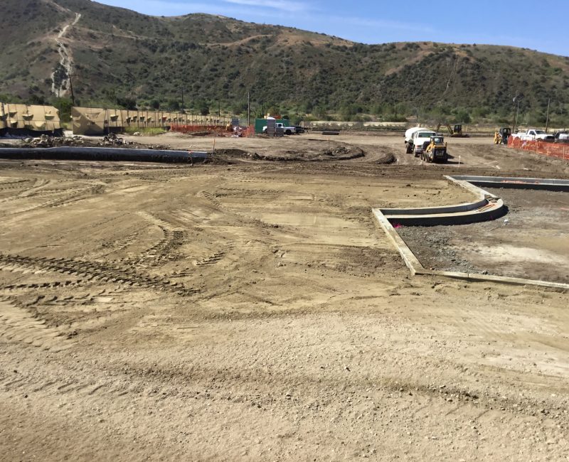 Construction site with dirt, vehicles, tire tracks, hills, clear sky.