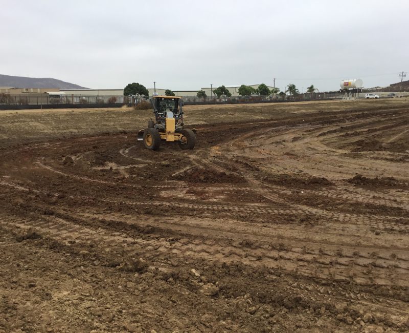 Construction vehicle on muddy field with buildings and cloudy sky.