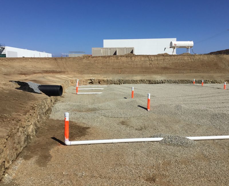 Gravel lot with pipes, orange markers, and buildings under blue sky.