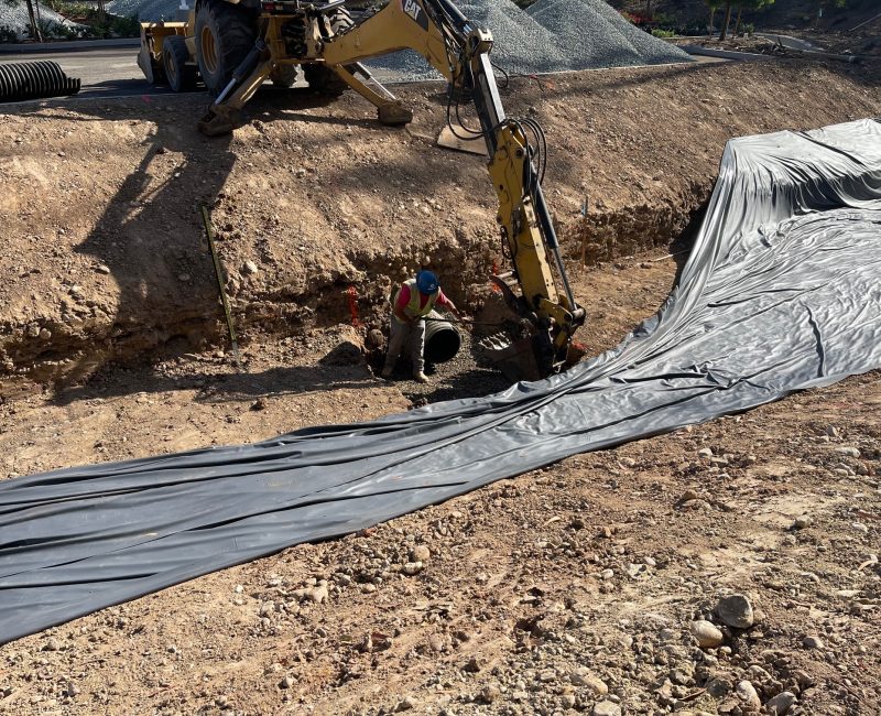 Workers use excavator to install black pipe in a trench.
