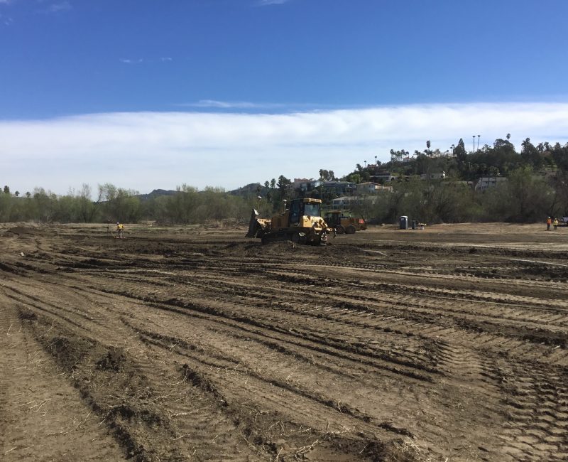 Bulldozer working on dirt, workers nearby, sky and houses visible.