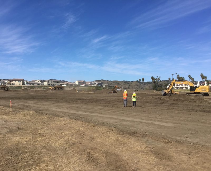 Two people in safety vests watch excavators at a construction site.