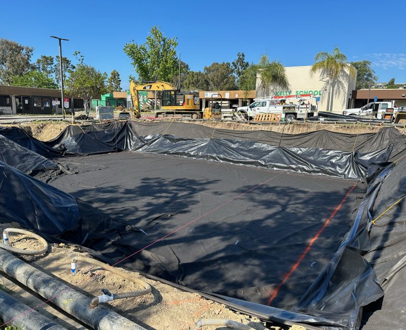 Construction site with lined pit, vehicles, and school in background.