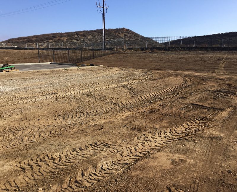 Dirt lot with tire tracks, gravel, fence, hills, blue sky.