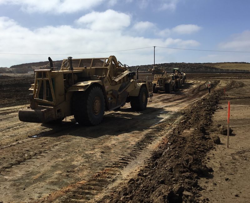 Earth-moving vehicles lined up on a dirt road, cloudy sky.