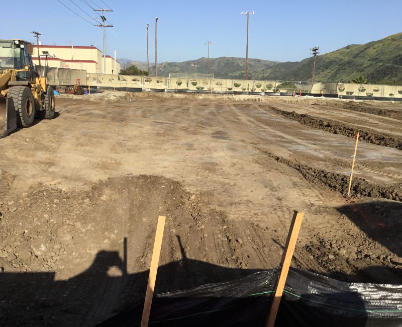Bulldozer, leveled dirt, stakes; hills and blue sky in background.