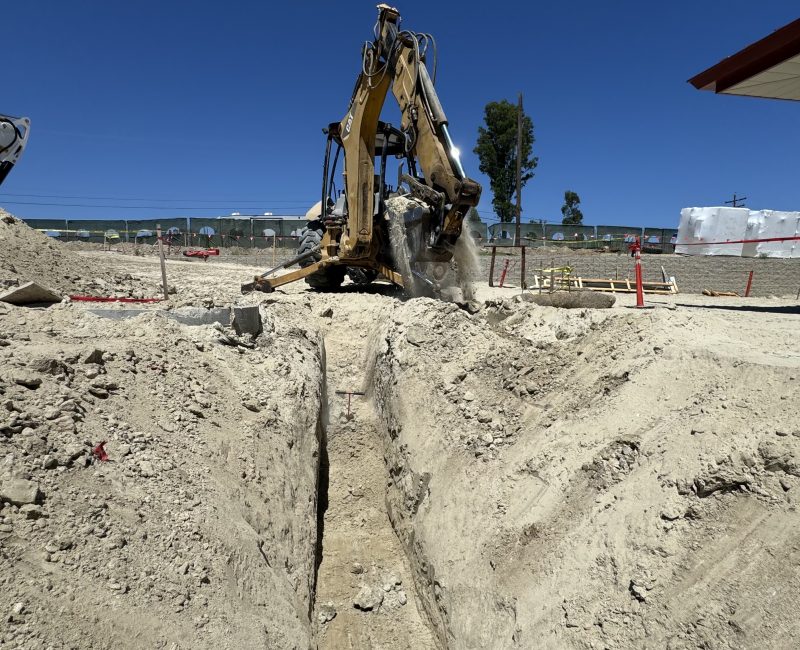 Yellow excavator digging at sunny construction site, dirt piles nearby.