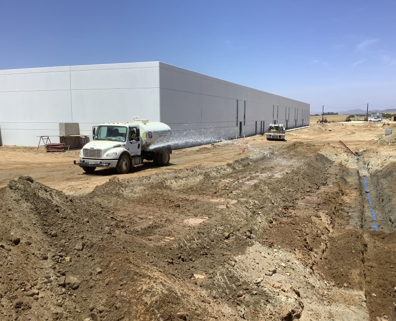 White water truck sprays dirt at construction site, blue sky overhead.