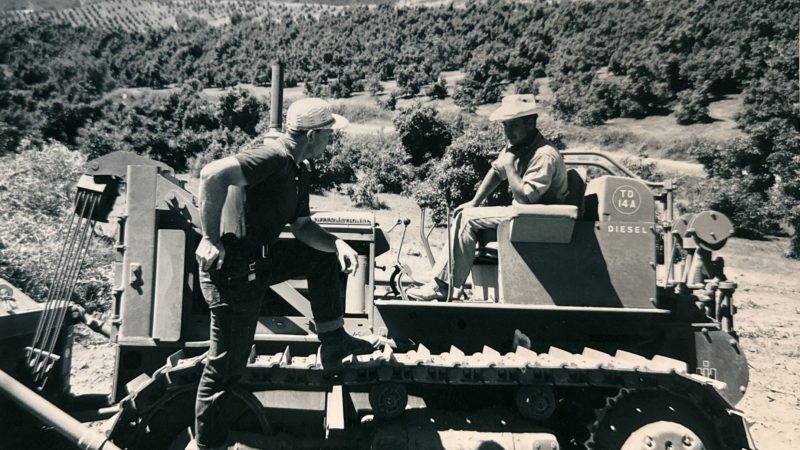 Two men by a diesel tractor in a hilly, rural landscape.