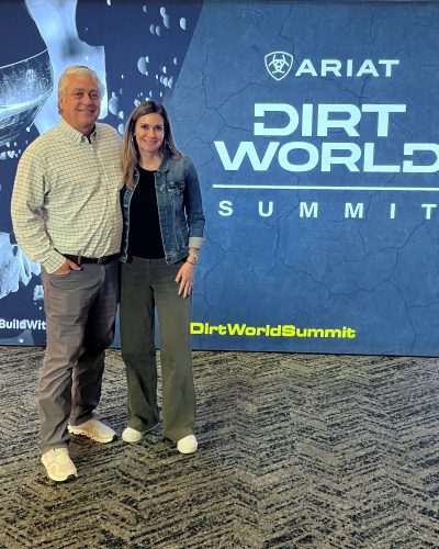 Two people smile before “ARIAT DIRT WORLD SUMMIT” industrial backdrop.