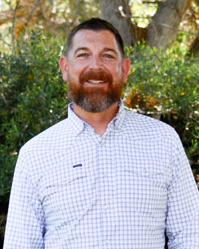 Bearded man in plaid shirt smiles outdoors by green foliage.