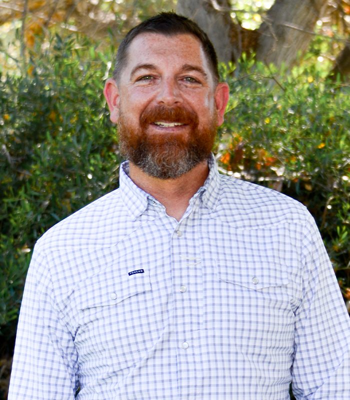 Bearded man in plaid shirt smiles outdoors by green foliage.