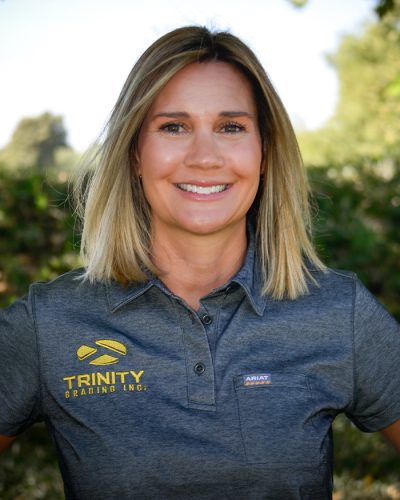 Blonde woman smiling outdoors in gray logo shirt, green trees behind.