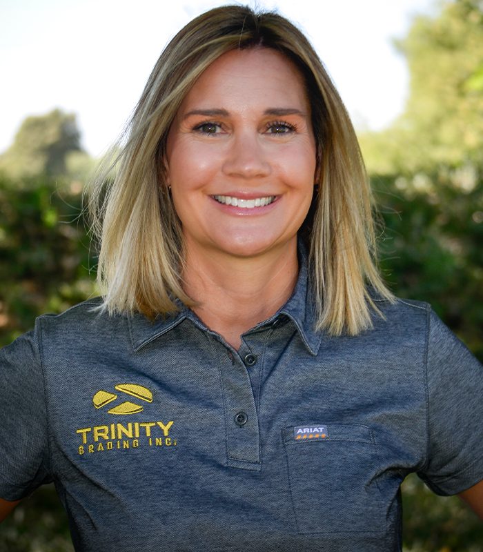 Blonde woman smiling outdoors in gray logo shirt, green trees behind.