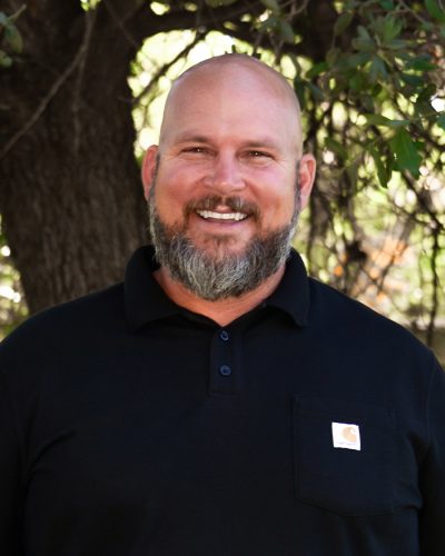 Bearded man in black shirt smiling by leafy tree outdoors.