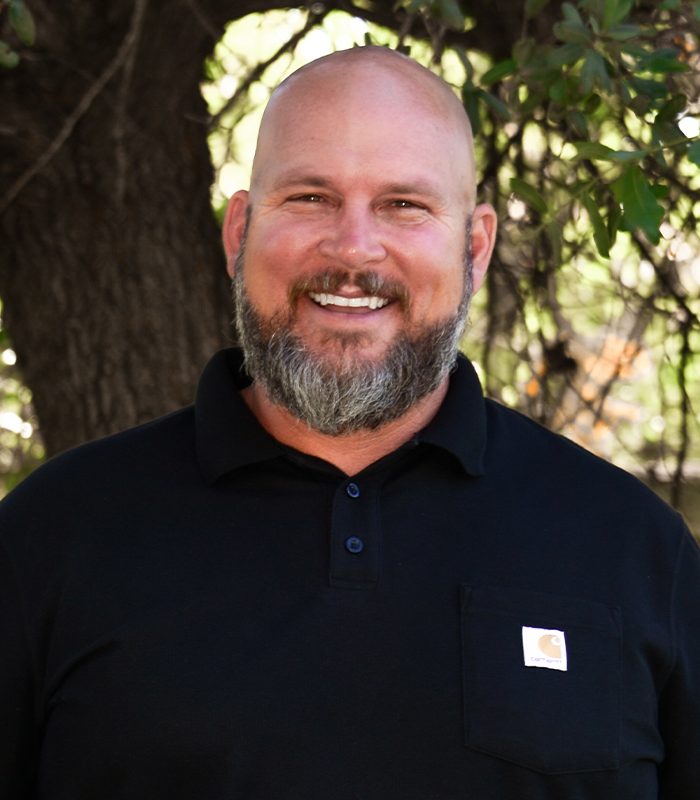 Bearded man in black shirt smiling by leafy tree outdoors.